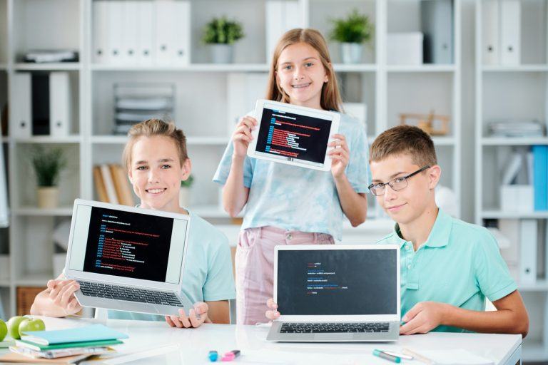 Three children proudly displaying their coding projects on laptops and tablets